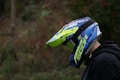 A person is wearing a bright, colorful motocross helmet with blue, green, and yellow accents. The helmet covers their face, and they are dressed in a dark jacket. The background is a blurred mix of natural colors, suggesting an outdoor setting.