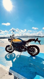 A Yamaha motorcycle is parked on a bright blue surface near a body of water. The sun is shining brightly in the sky, casting a clear shadow of the motorcycle. Fluffy white clouds are scattered across the blue sky, and the landscape in the background appears to be hilly.