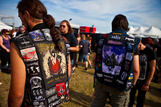 Several people are gathered outdoors at a festival or concert. Two individuals in the foreground are wearing denim jackets adorned with multiple patches of different bands and designs. The background shows a crowd of people and large white tents set up under a blue sky.