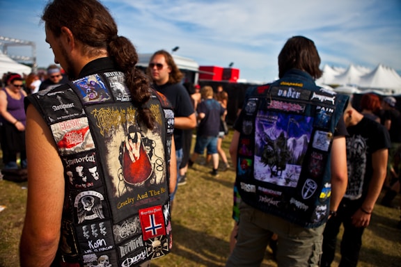 Several people are gathered outdoors at a festival or concert. Two individuals in the foreground are wearing denim jackets adorned with multiple patches of different bands and designs. The background shows a crowd of people and large white tents set up under a blue sky.