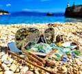 Sunglasses rest on a pebble beach surrounded by colorful sea glass and driftwood. The lenses reflect part of the beach environment. The clear blue sea and a distant cliff with two figures are visible in the background.