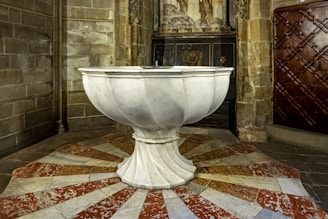 An ornate, white marble baptismal font is situated on a circular patterned tile floor with red and white sections. The surrounding area features stone walls and a carved wooden door with geometric designs. In the background, there is an elaborate relief carving, possibly depicting religious figures.