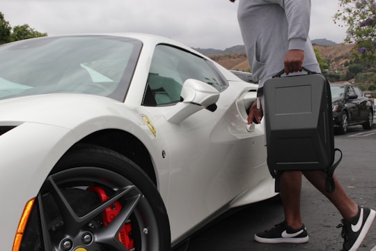 A person holding a black case is unlocking a sleek, white sports car in a parking lot. The car has red brake calipers and distinct wheel designs. The surrounding environment includes overcast skies and distant hills with sparse vegetation.