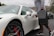 A person holding a black case is unlocking a sleek, white sports car in a parking lot. The car has red brake calipers and distinct wheel designs. The surrounding environment includes overcast skies and distant hills with sparse vegetation.