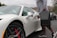 A person holding a black case is unlocking a sleek, white sports car in a parking lot. The car has red brake calipers and distinct wheel designs. The surrounding environment includes overcast skies and distant hills with sparse vegetation.