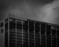 A tall building with a modern glass facade stands against a moody, overcast sky. The building displays the name 'Sheraton Maria Isabel' near the top. The windows are mostly dark, with a few lights on inside.