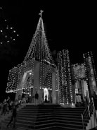 A night scene featuring a church adorned with strings of lights that illuminate its facade and spire. A cross tops the spire, and people stand and sit on the church steps. The scene has a festive and serene atmosphere.