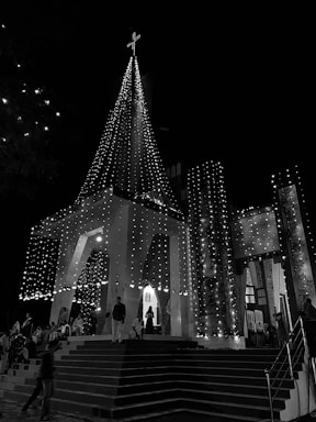 A night scene featuring a church adorned with strings of lights that illuminate its facade and spire. A cross tops the spire, and people stand and sit on the church steps. The scene has a festive and serene atmosphere.