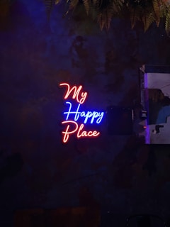Neon lights form the words 'My Happy Place' against a dark, textured wall with hanging ferns above. The letters are in vibrant colors, with 'My' and 'Place' in red, and 'Happy' in blue.