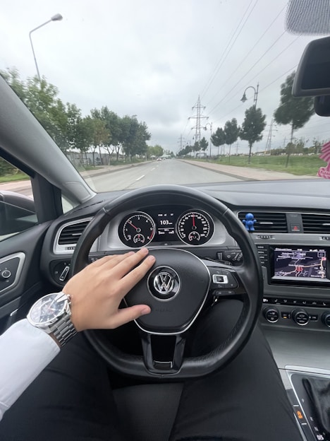 Interior view of a car showing the driver's perspective with a hand on the steering wheel featuring the Volkswagen logo. The dashboard displays the speedometer, and a GPS screen is visible. The road is lined with trees and streetlights, under a cloudy sky.