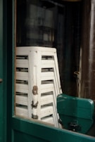 A stack of white stools with peeling paint are positioned behind a green-framed window. The stools are slightly worn, showing signs of rust and aging on the metal components.