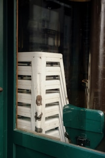 A stack of white stools with peeling paint are positioned behind a green-framed window. The stools are slightly worn, showing signs of rust and aging on the metal components.