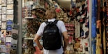 A person with a backpack is standing in front of a souvenir shop. The shop is filled with various items including hats, t-shirts, keychains, and postcards. On the left, there is a display of postcards, while on the right, shelves are stocked with numerous souvenir items. The mood is vibrant and busy, typical of tourist areas.