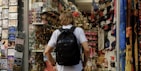 A person with a backpack is standing in front of a souvenir shop. The shop is filled with various items including hats, t-shirts, keychains, and postcards. On the left, there is a display of postcards, while on the right, shelves are stocked with numerous souvenir items. The mood is vibrant and busy, typical of tourist areas.