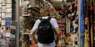 A person with a backpack is standing in front of a souvenir shop. The shop is filled with various items including hats, t-shirts, keychains, and postcards. On the left, there is a display of postcards, while on the right, shelves are stocked with numerous souvenir items. The mood is vibrant and busy, typical of tourist areas.