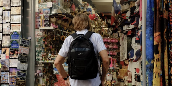 A person with a backpack is standing in front of a souvenir shop. The shop is filled with various items including hats, t-shirts, keychains, and postcards. On the left, there is a display of postcards, while on the right, shelves are stocked with numerous souvenir items. The mood is vibrant and busy, typical of tourist areas.