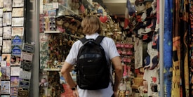 A person with a backpack is standing in front of a souvenir shop. The shop is filled with various items including hats, t-shirts, keychains, and postcards. On the left, there is a display of postcards, while on the right, shelves are stocked with numerous souvenir items. The mood is vibrant and busy, typical of tourist areas.