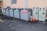 A series of recycling bins lined up against a beige wall, each labeled for different types of materials like brown glass, green glass, and aluminum. Some bins have waste sticking out, indicating they are full. Nearby, a few bags and loose pieces of litter are scattered on the ground.