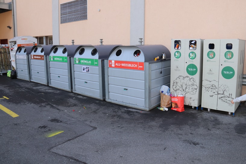 A series of recycling bins lined up against a beige wall, each labeled for different types of materials like brown glass, green glass, and aluminum. Some bins have waste sticking out, indicating they are full. Nearby, a few bags and loose pieces of litter are scattered on the ground.