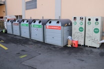 A series of recycling bins lined up against a beige wall, each labeled for different types of materials like brown glass, green glass, and aluminum. Some bins have waste sticking out, indicating they are full. Nearby, a few bags and loose pieces of litter are scattered on the ground.
