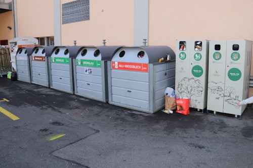 A series of recycling bins lined up against a beige wall, each labeled for different types of materials like brown glass, green glass, and aluminum. Some bins have waste sticking out, indicating they are full. Nearby, a few bags and loose pieces of litter are scattered on the ground.