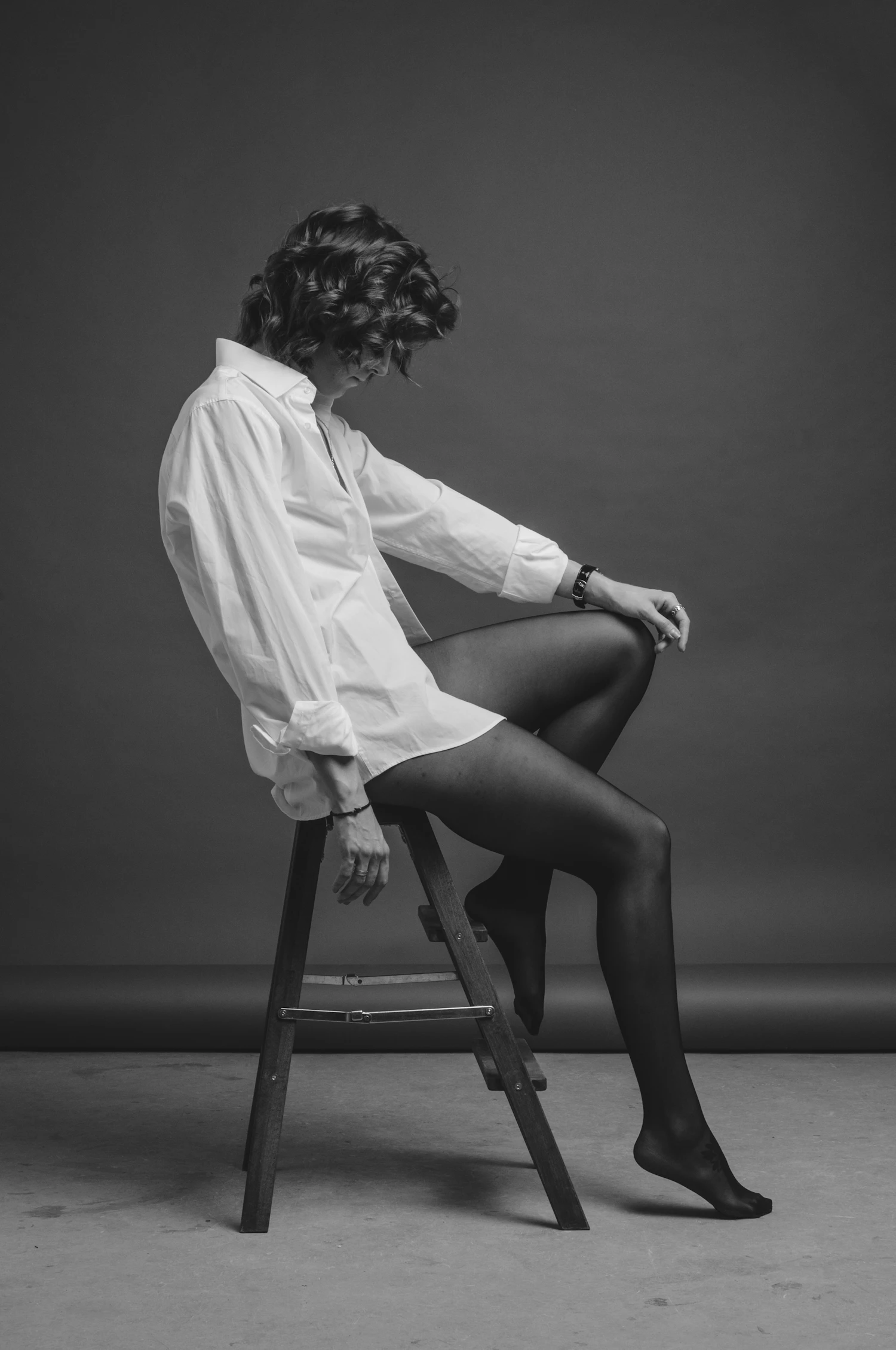 A woman seated on a wooden stool in a studio, full length, one arm across her knee