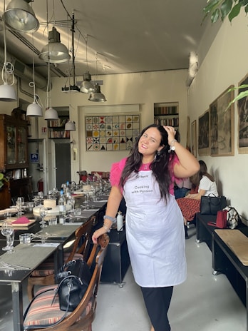 A woman in a pink shirt and white apron is standing and smiling in a dining room setting with tables prepared for a meal. The room features modern hanging lights, decorative wall art, and wooden chairs. There are glasses, napkins, and tableware neatly arranged on the tables.