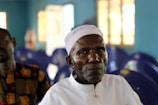 An elderly man wearing a white cap and white shirt is sitting in a room with blue chairs and walls. The atmosphere appears peaceful, with soft lighting coming from windows in the background. Another person is partially visible but out of focus.