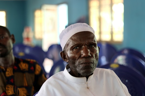 An elderly man wearing a white cap and white shirt is sitting in a room with blue chairs and walls. The atmosphere appears peaceful, with soft lighting coming from windows in the background. Another person is partially visible but out of focus.