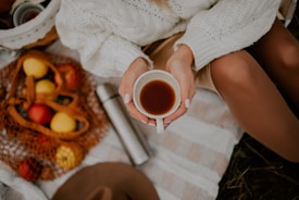 A person in a cozy knitted sweater is holding a cup of hot beverage, surrounded by picnic items including a thermos and a mesh bag filled with various fruits on a checkered blanket.