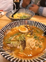 Two bowls of ramen are placed on a wooden table. Each bowl contains noodles submerged in a rich, flavorful broth, topped with ingredients like boiled eggs, sliced green onions, corn, and meat. A person can be seen partially in the background, suggesting a dining environment.