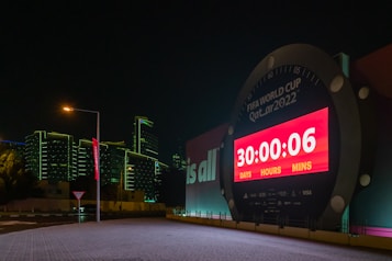 A large countdown clock displays the time left until the FIFA World Cup Qatar 2022. The digital timer shows '30 days, 0 hours, and 6 minutes' remaining. The clock is illuminated by bright red and white lights and is set against a backdrop of modern buildings with green outline lighting at night. A streetlamp and a road sign are visible in the foreground, adding to the urban setting.