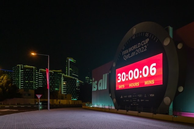 A large countdown clock displays the time left until the FIFA World Cup Qatar 2022. The digital timer shows '30 days, 0 hours, and 6 minutes' remaining. The clock is illuminated by bright red and white lights and is set against a backdrop of modern buildings with green outline lighting at night. A streetlamp and a road sign are visible in the foreground, adding to the urban setting.