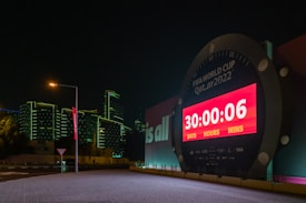 A large countdown clock displays the time left until the FIFA World Cup Qatar 2022. The digital timer shows '30 days, 0 hours, and 6 minutes' remaining. The clock is illuminated by bright red and white lights and is set against a backdrop of modern buildings with green outline lighting at night. A streetlamp and a road sign are visible in the foreground, adding to the urban setting.