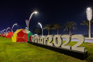 Large 3D letters spelling out '#Qatar2022' are prominently displayed on a grassy area beside a road, illuminated by modern street lamps. In the background, colorful geometric structures resembling giant soccer balls are lined up, and palm trees are visible under a dark night sky.