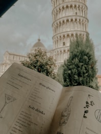 A menu is prominently displayed in the foreground, with appetizers, wine glasses, and soft drinks listed. In the background, the Leaning Tower of Pisa is visible, partially obscured by tall trees and a cloudy sky.
