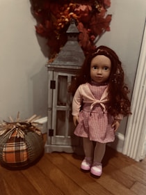 A doll with long brown hair stands next to a decorative lantern on a wooden floor. The doll is dressed in a red and white checkered dress with a pink cardigan and pink shoes. Beside the doll, there is a fabric pumpkin with plaid patterns, and autumn leaves are visible behind the lantern.