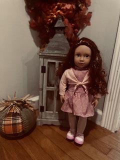 A doll with long brown hair stands next to a decorative lantern on a wooden floor. The doll is dressed in a red and white checkered dress with a pink cardigan and pink shoes. Beside the doll, there is a fabric pumpkin with plaid patterns, and autumn leaves are visible behind the lantern.