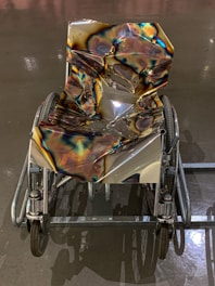 A wheelchair with a highly reflective, metallic surface that appears warped and distorted, coated with iridescent, rainbow-like patterns. The floor beneath is smooth and reflective, possibly a polished concrete surface, enhancing the visual effect.