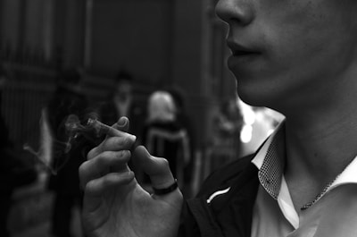 A close-up image of a person holding a lit cigarette, with smoke visibly swirling in the air. The person's lips and part of their face are partially visible, while background figures appear blurred and out of focus, giving a sense of urban environment.