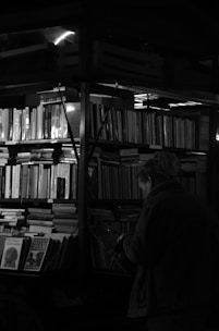 A dimly lit scene featuring a person browsing a bookshelf filled with numerous books. The environment suggests an indoor or possibly an outdoor bookstall. Shelves are tightly packed with books, some of which have visible titles. The lighting is subdued, casting shadows and highlighting the contours of the person's silhouette.