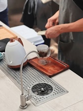 A person wearing an apron appears to be preparing a beverage at a countertop. A ceramic kettle with a wooden handle rests beside a clear glass cup containing tea leaves, placed on a wooden tray. A stainless steel faucet and drain are visible in the foreground.