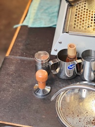 A coffee preparation area featuring a tamper with a wooden handle, a metal shaker, a milk frothing jug, and an espresso machine tray with a blue cloth nearby. Coffee grounds are scattered on the surface near a metallic sink.
