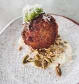 A round, crispy-fried food item topped with a dollop of green sauce and shredded white cheese is placed on a textured plate. It sits on a bed of assorted seeds and nuts with a creamy white sauce underneath.