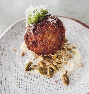 A round, crispy-fried food item topped with a dollop of green sauce and shredded white cheese is placed on a textured plate. It sits on a bed of assorted seeds and nuts with a creamy white sauce underneath.