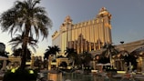 A grand, ornate building with golden illumination stands prominently, reflecting the evening light. Palm trees surround a luxurious pool area, creating an opulent resort atmosphere. The architecture features intricate details and a blend of classic and modern design elements.