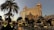 A grand, ornate building with golden illumination stands prominently, reflecting the evening light. Palm trees surround a luxurious pool area, creating an opulent resort atmosphere. The architecture features intricate details and a blend of classic and modern design elements.