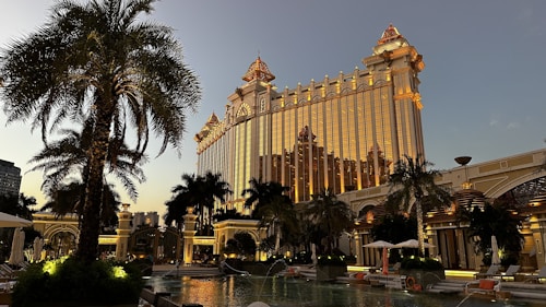 A grand, ornate building with golden illumination stands prominently, reflecting the evening light. Palm trees surround a luxurious pool area, creating an opulent resort atmosphere. The architecture features intricate details and a blend of classic and modern design elements.