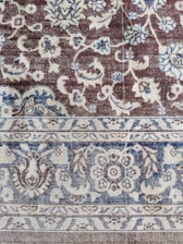 An intricately woven carpet with floral and geometric patterns. The design includes flowers, leaves, and symmetric motifs in blue and beige colors set against a brown background.