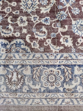 An intricately woven carpet with floral and geometric patterns. The design includes flowers, leaves, and symmetric motifs in blue and beige colors set against a brown background.